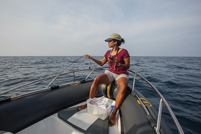 Asha de Vos is sitting on a boat, holding laboratory equipment in both hands. She is wearing a baseball cap, a maroon T-shirt and a pair of white shorts. She is not wearing shoes.